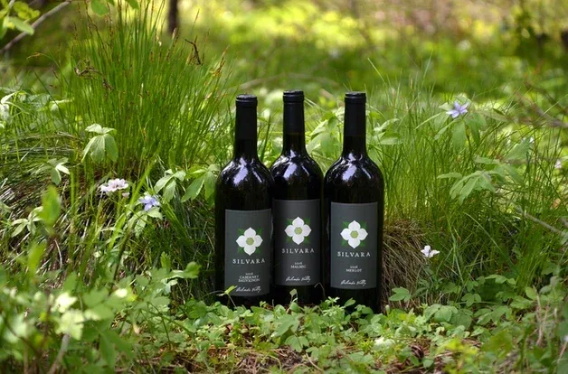 Three dark wine bottles labeled Silvara placed on the ground among green grass and small purple flowers in a natural outdoor setting.