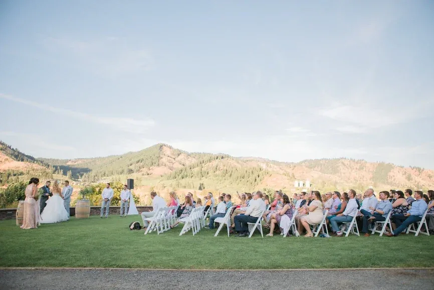 Outdoor wedding ceremony at Silvara Cellars in Leavenworth with bridal party and guests seated on white chairs against a backdrop of rolling hills under a clear sky.