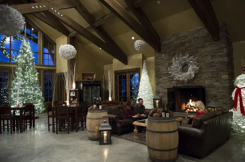 Cozy indoor scene with three people seated on brown leather sofas around a wooden coffee table, next to a stone fireplace with a burning fire, decorated with Christmas trees, a snowman, wreaths, and hanging ornaments.