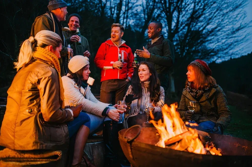 A group of friends wearing warm clothing relax around a campfire in the evening outdoors, holding drinks and engaging in lively conversation.
