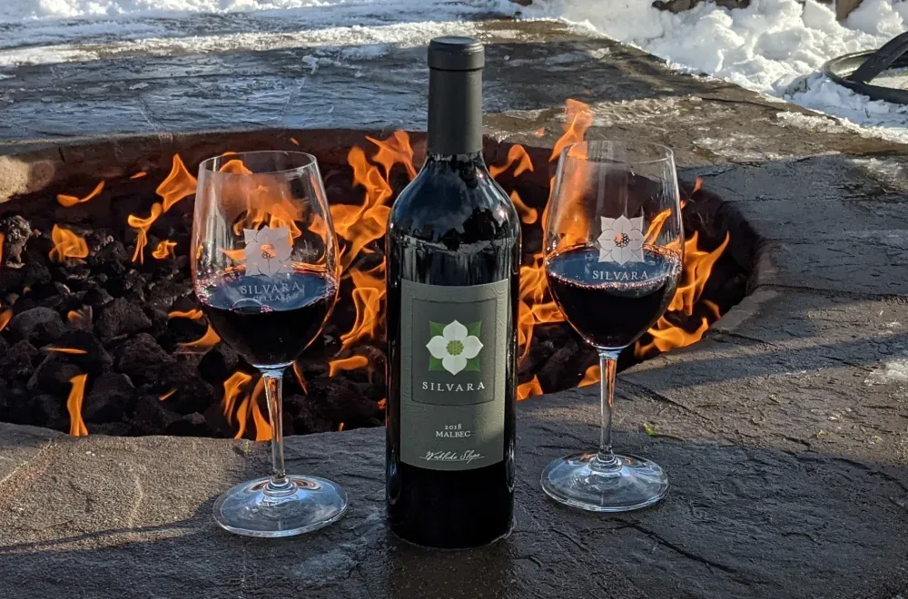 A bottle of Silvara Malbec wine flanked by two filled wine glasses set in front of a roaring outdoor fire pit on a stone surface with snow in the background.