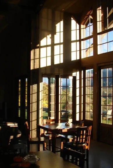 Sunlight streaming through large windows casting shadows around a wooden table and chairs in a cozy wine tasting room with a scenic view outside.