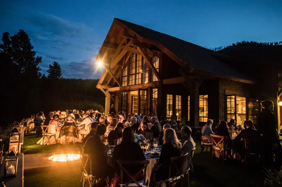 Evening outdoor gathering at a warmly lit rustic wooden building with large windows and a pitched roof, where guests are dining around tables with a glowing fire pit nearby and surrounded by trees under a darkening sky.