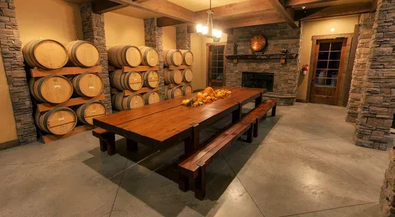 Wine tasting room with wooden barrels stacked against stone walls, a long wooden table with benches, and soft overhead lighting in a rustic setting.
