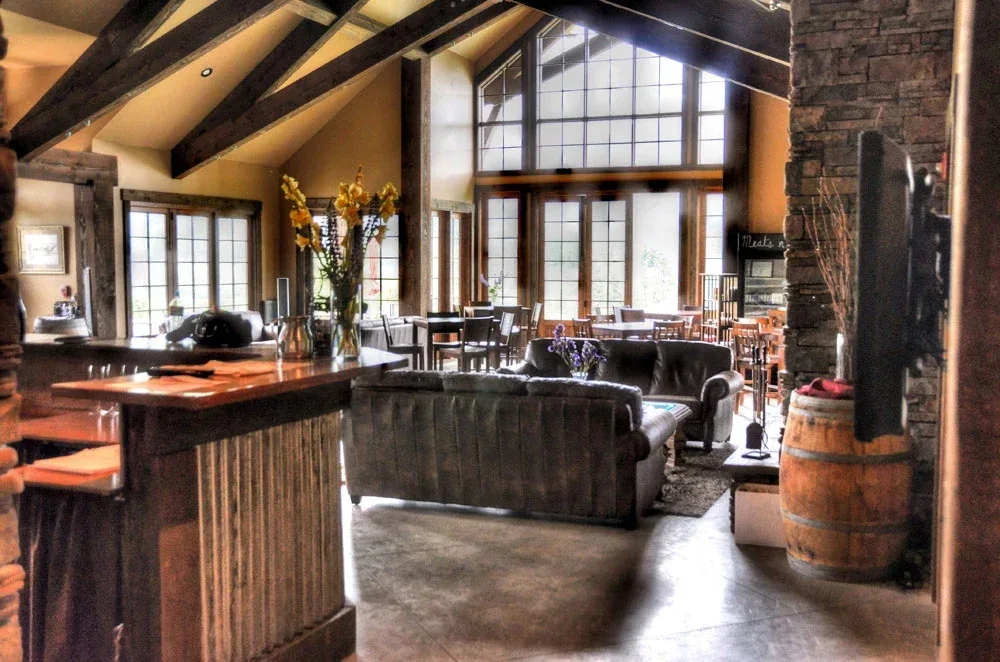 Cozy indoor wine tasting room with wooden beams, leather sofas, large windows letting in natural light, a bar counter with flowers, and rustic decor including a wine barrel and stone wall.