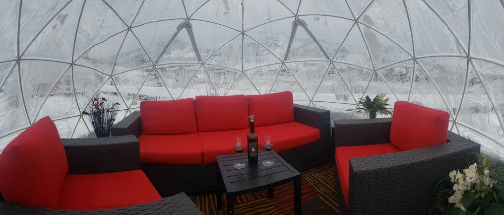 Interior of an igloo-shaped tasting room with black wicker furniture featuring bright red cushions, a small black table with a wine bottle and two glasses, surrounded by snow-covered windows.