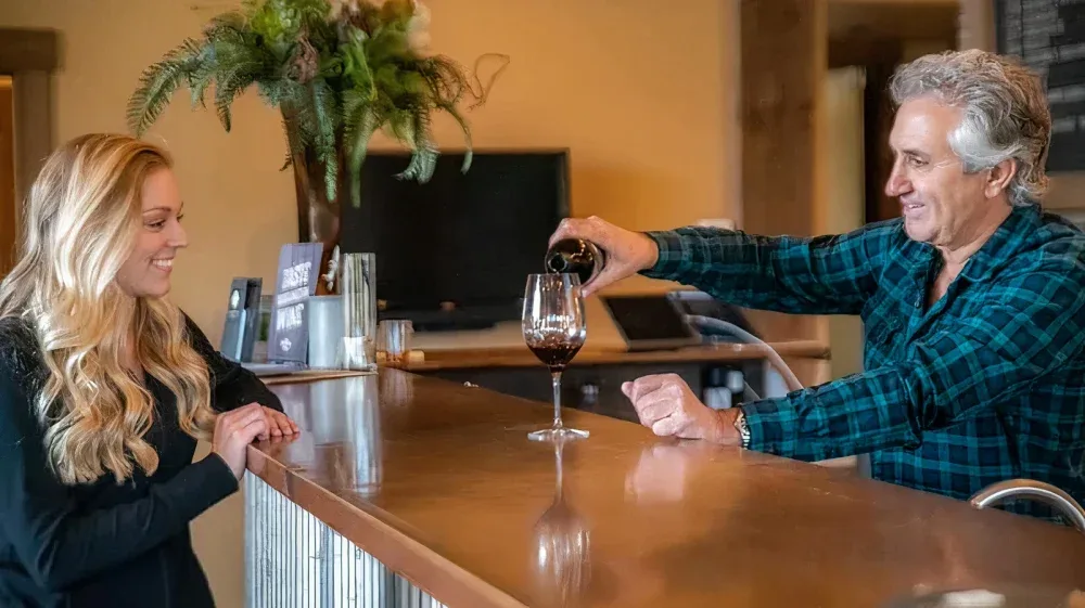 A man behind a bar in a tasting room pours red wine into a glass while a smiling woman watches from the other side of the counter. The setting is cozy with a floral arrangement and a yellow wall in the background.