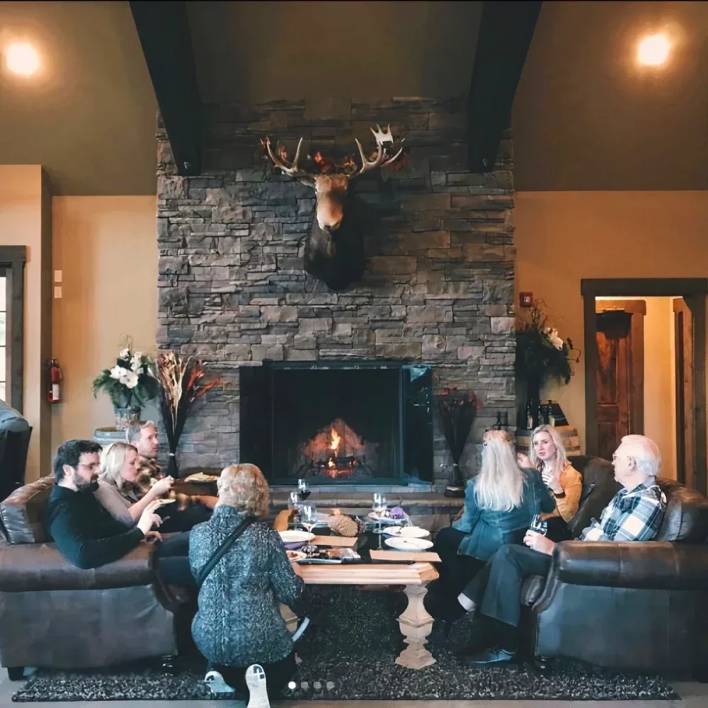 A group of people seated on sofas around a wooden table in a cozy tasting room with a stone fireplace and a mounted deer head above it in Leavenworth.
