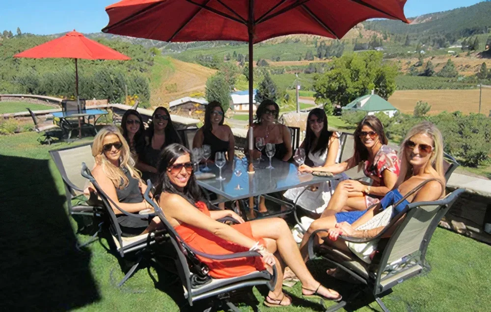 A group of nine women wearing sunglasses seated around a glass table with empty wine glasses on a sunny terrace overlooking green hills and farmland, shaded by red umbrellas.