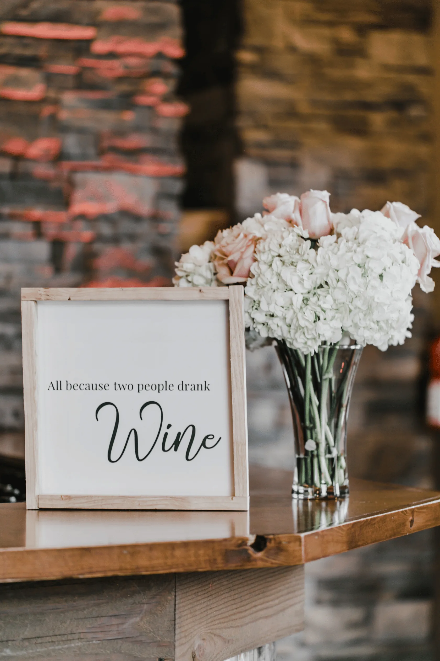 Wooden sign reading “All because two people drank wine” beside roses and hydrangeas on a counter at Silvara Cellars