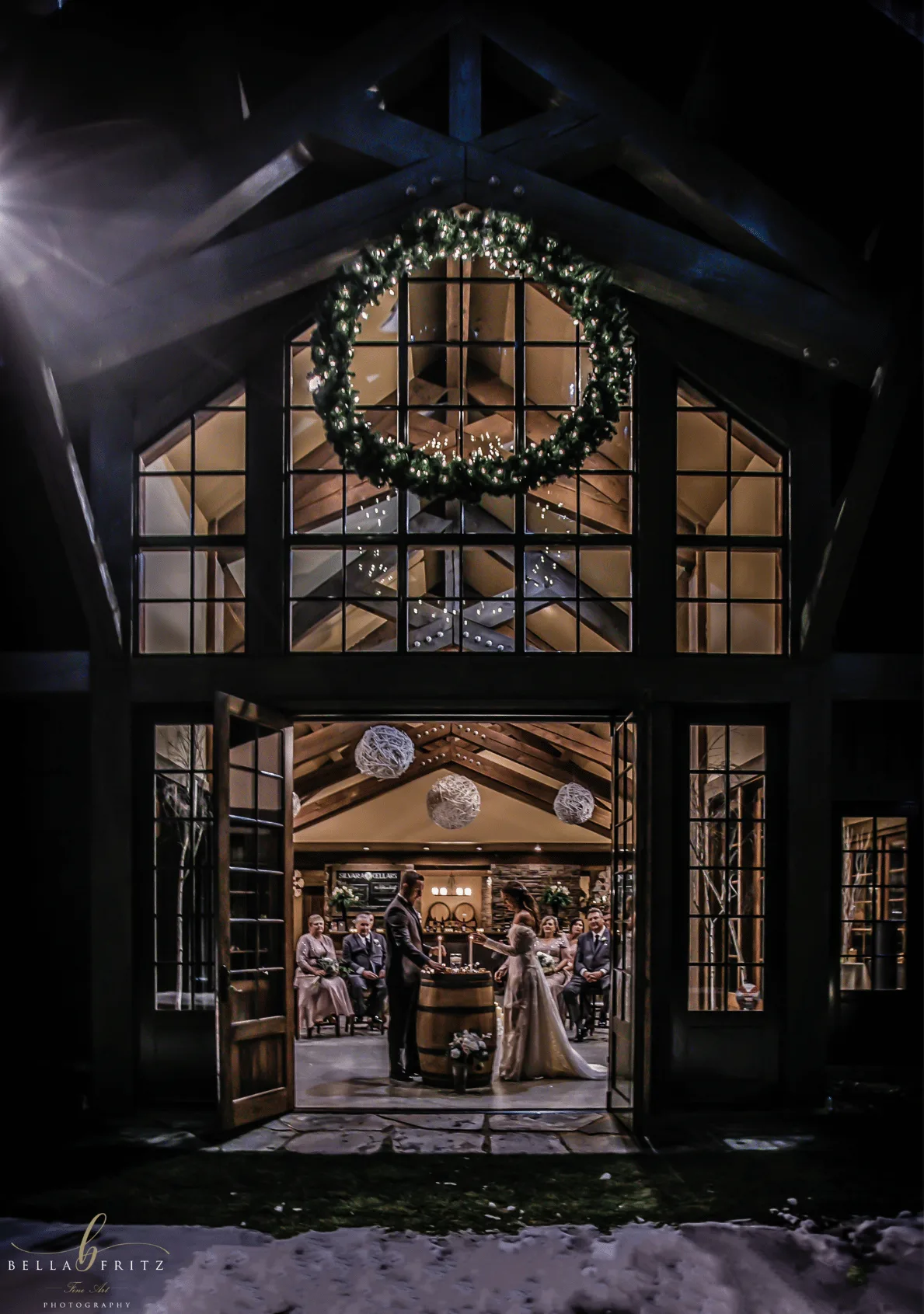 Couple marrying inside Silvara Cellars on a winter night, seen through the open doors beneath a lit wreath