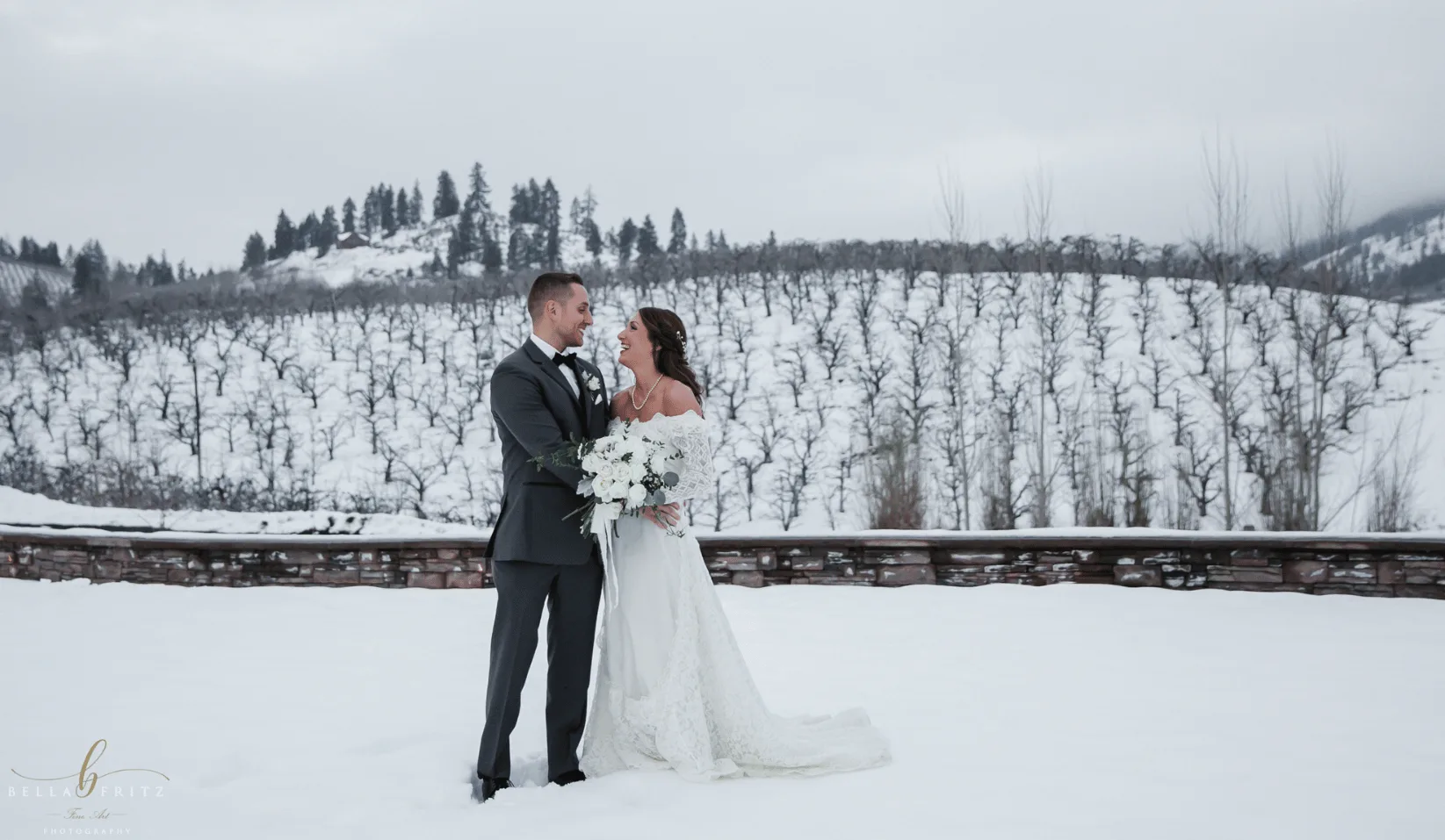 Bride and groom standing in deep snow at Silvara Cellars, with a snow-covered orchard on the hillside behind them