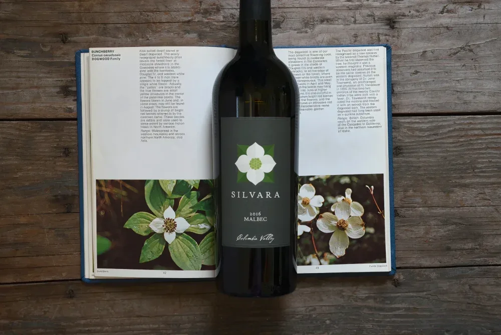 A bottle of Silvara 2016 Malbec wine placed on top of an open book featuring illustrations and descriptions of dogwood flowers, set on a wooden surface.