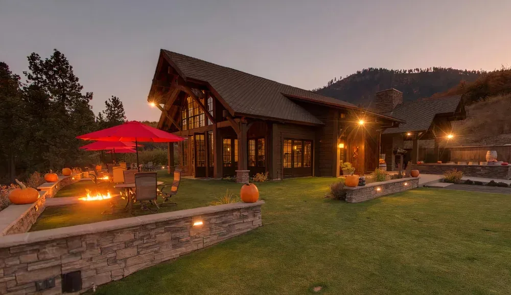 A rustic wooden house at dusk with outdoor seating featuring a fire pit and red umbrellas, surrounded by pumpkins and illuminated by warm lights in a landscaped yard with hills in the background.