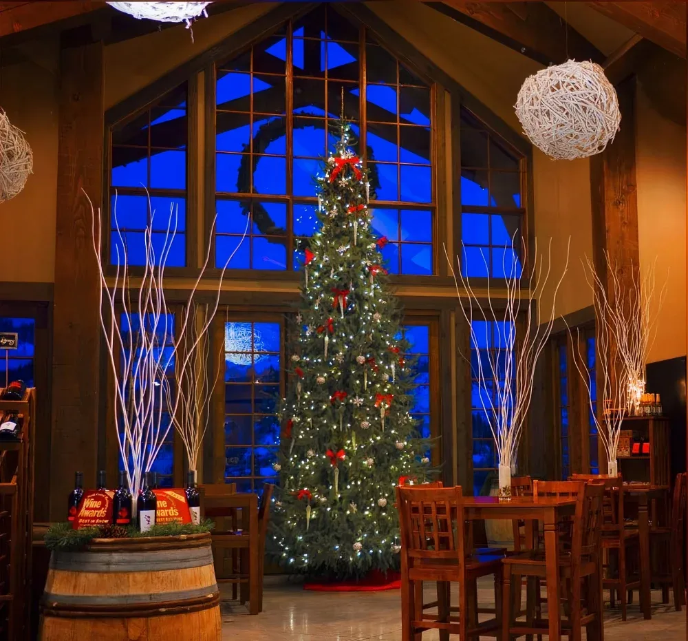 Tall, decorated Christmas tree with white lights and red bows in a warmly lit tasting room with wooden furniture and large windows showing a deep blue evening sky.