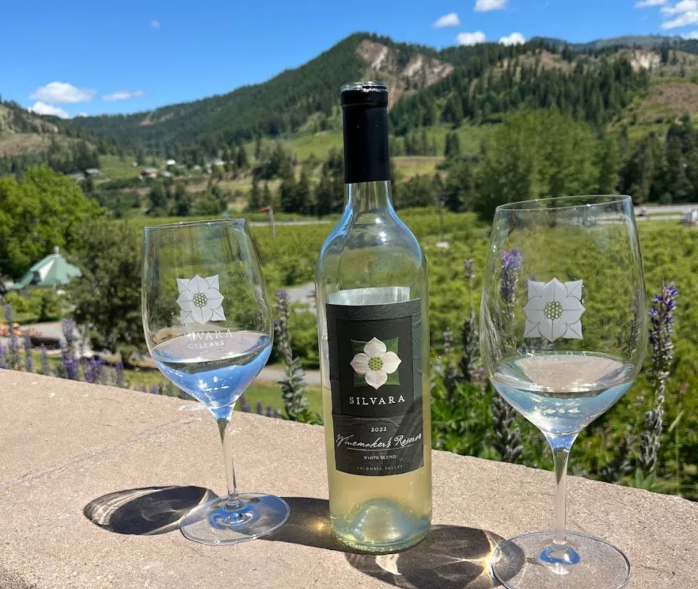 A bottle of Silvara Cellars 2022 Winemaker's Reserve white wine flanked by two wine glasses containing white wine on an outdoor table with a scenic view of greenery and hills in Leavenworth, Washington.