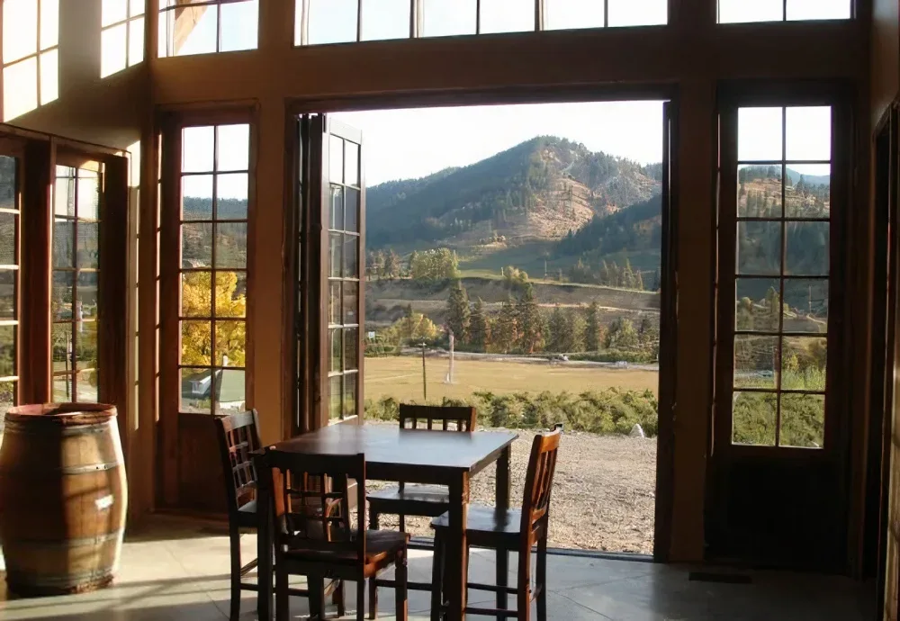 A wine tasting room with wooden chairs and a table near open French doors revealing a scenic view of a grassy field and forested hills under bright daylight.