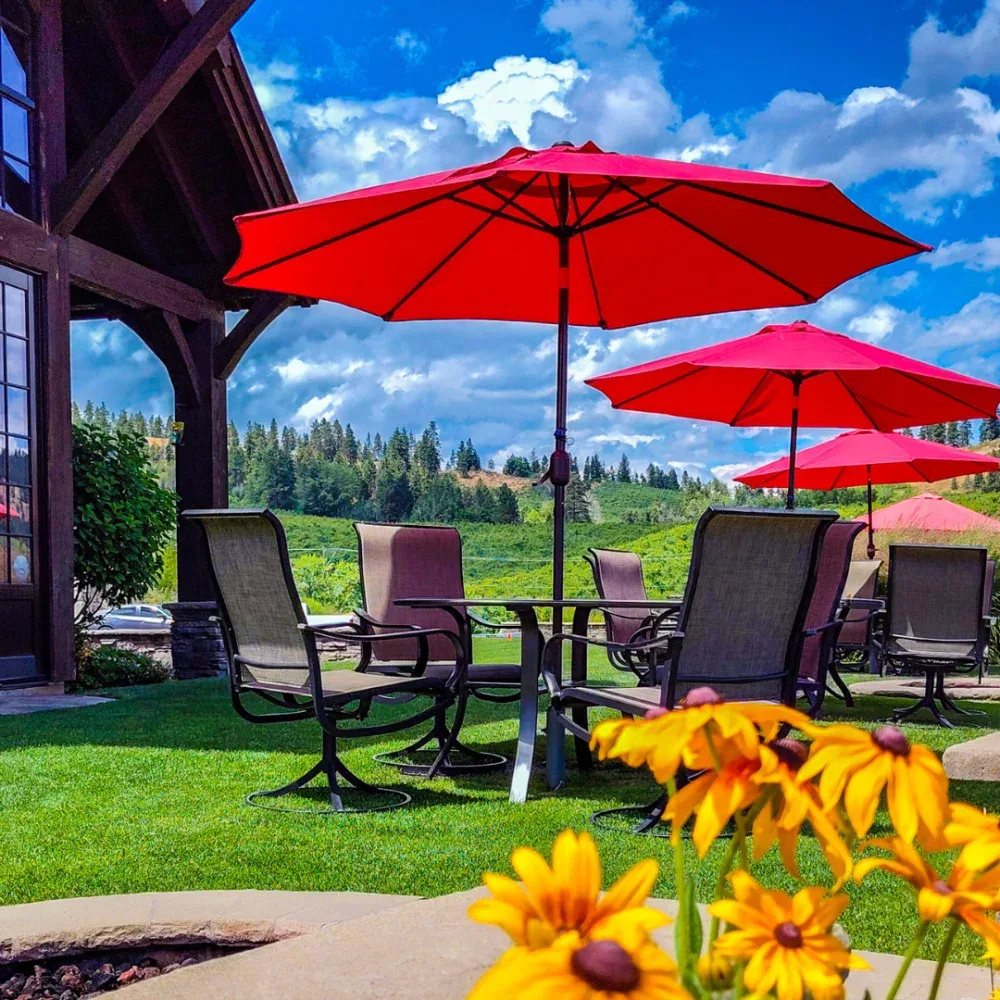 Outdoor patio seating area at a tasting room with several red umbrellas shading brown chairs and tables, surrounded by green grass and flowers, with a forested hill in the background under a partly cloudy sky.