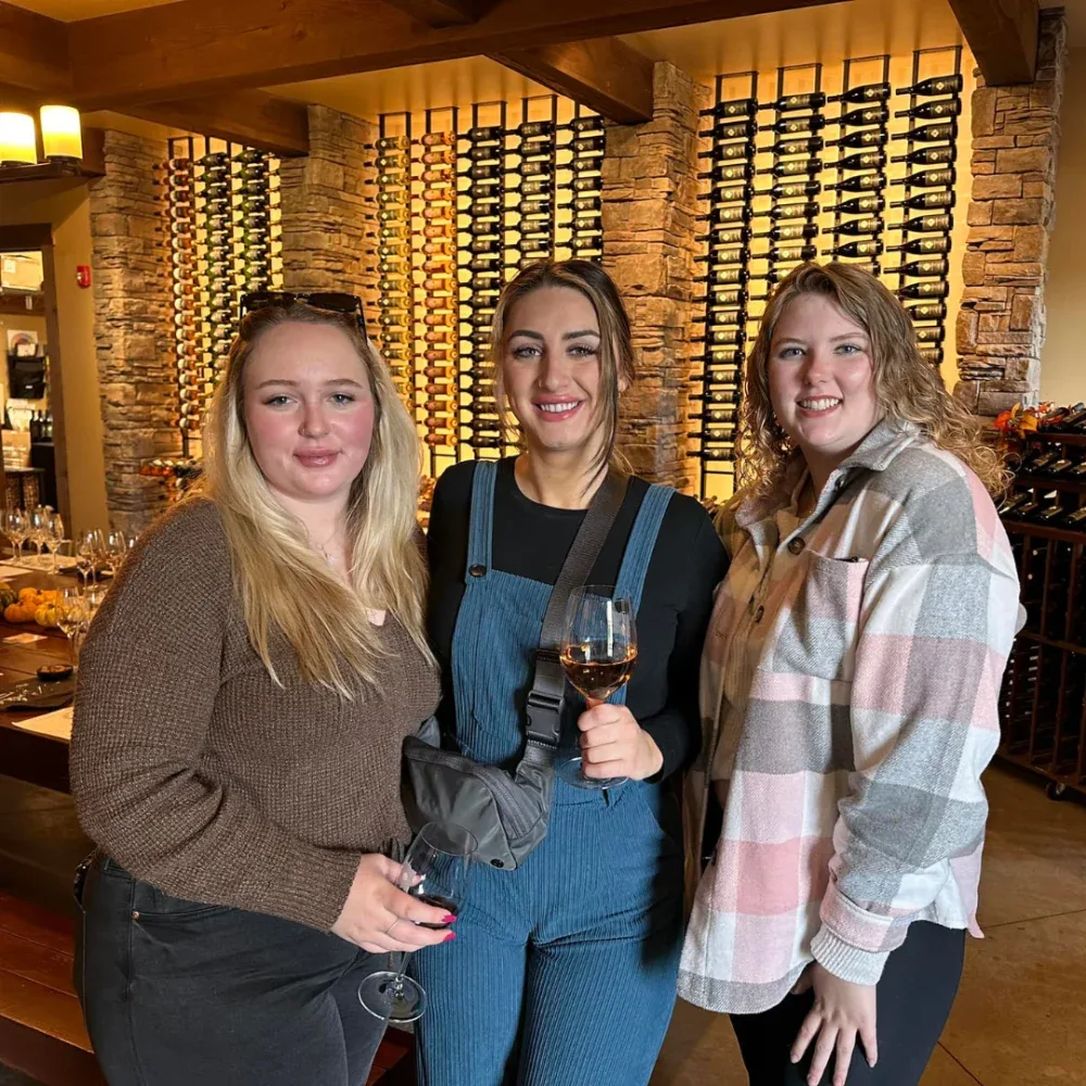 Three women smiling and posing for a photo inside a wine tasting room with wine bottles displayed on stone walls behind them. One woman in the center is holding a glass of rosé wine.