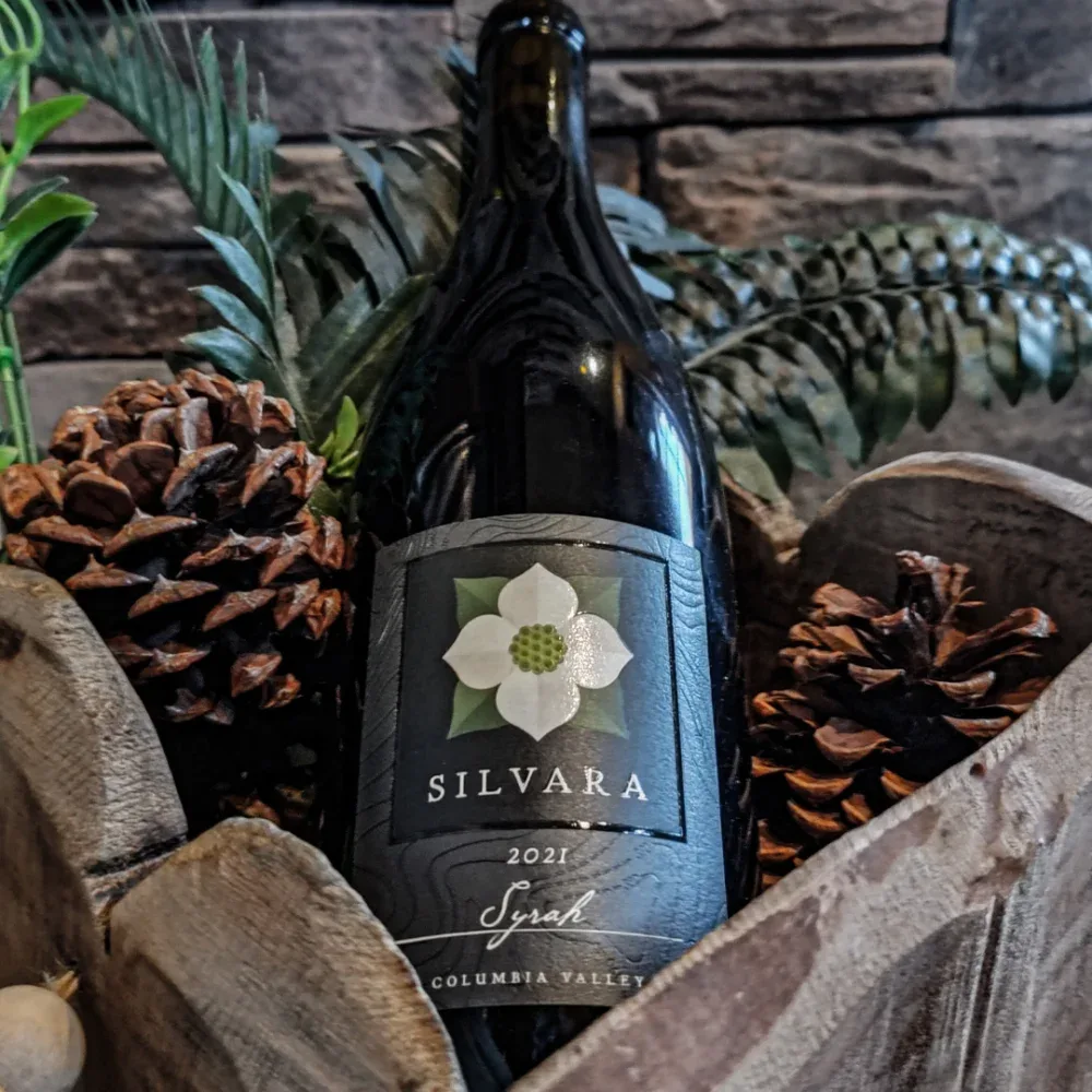 A close-up of a Silvara wine bottle from 2021 surrounded by pine cones and green foliage inside a wooden container, with a rustic background.