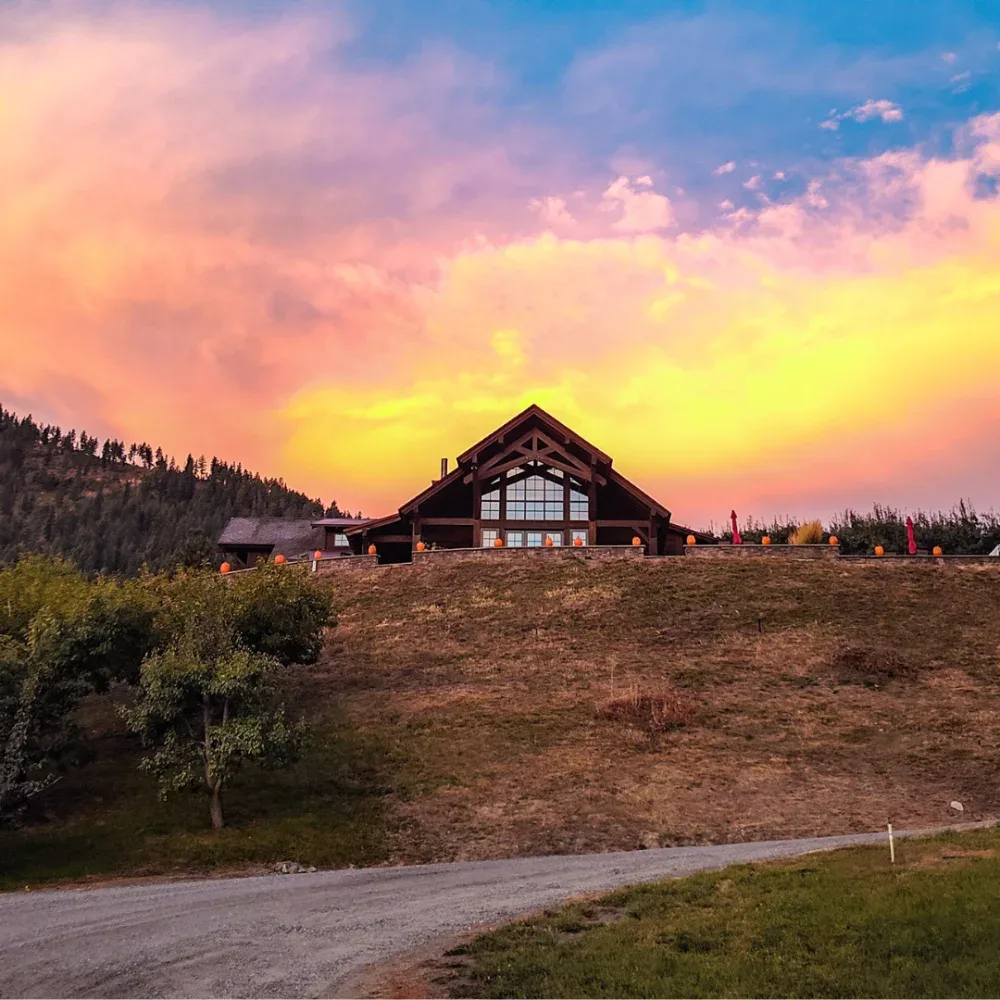 A rustic wooden building with large windows on a hill, set against a vibrant yellow and pink sky at sunset, surrounded by trees and forested mountains.