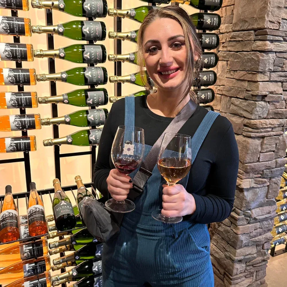 Smiling woman in a black shirt and blue overalls holds two wine glasses with red and white wine inside Silvara Cellars tasting room, with shelves of wine bottles and a stone wall in the background.
